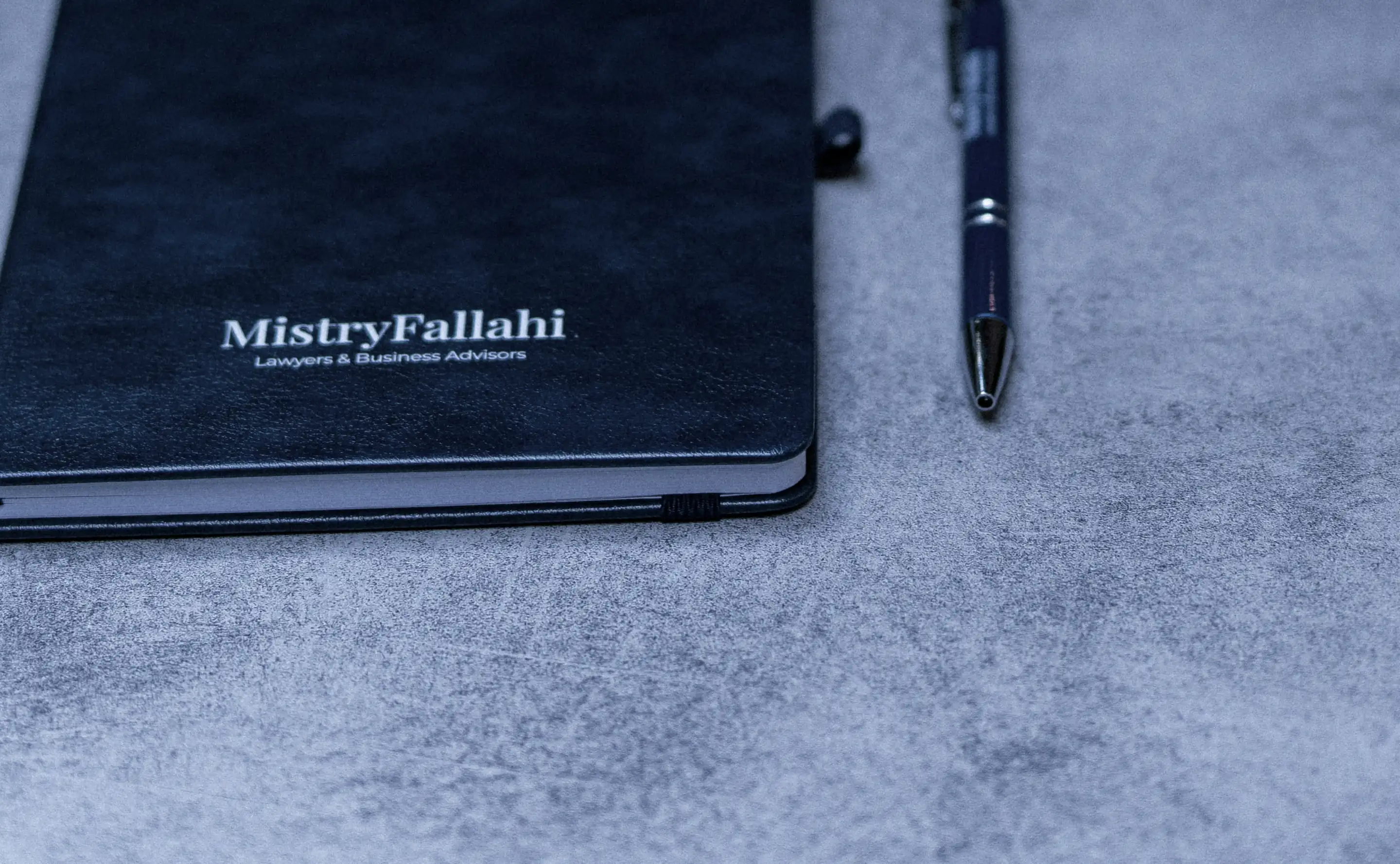 A navy MistryFallahi notebook and pen on a grey table, with a coffee beside them
