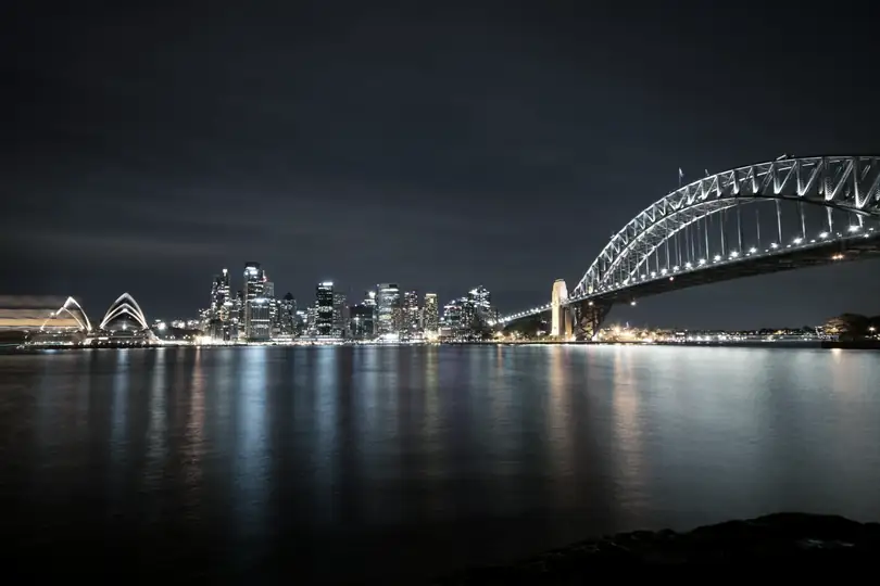 A harbour at night with a lit city skyline and an arch bridge