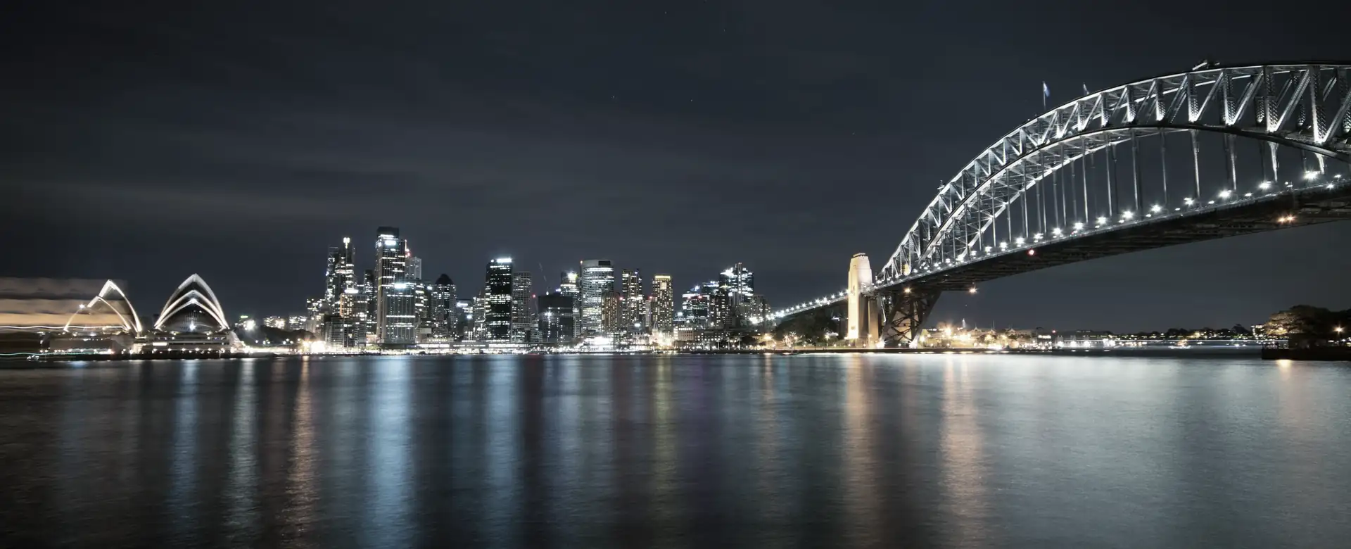 A harbour at night with a lit city skyline and an arch bridge