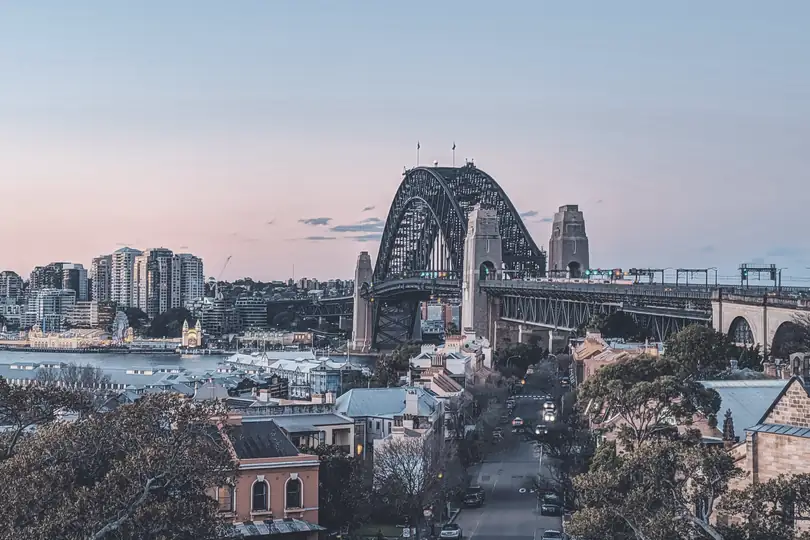A harbour bridge and city towers at dusk, seen over rooftops