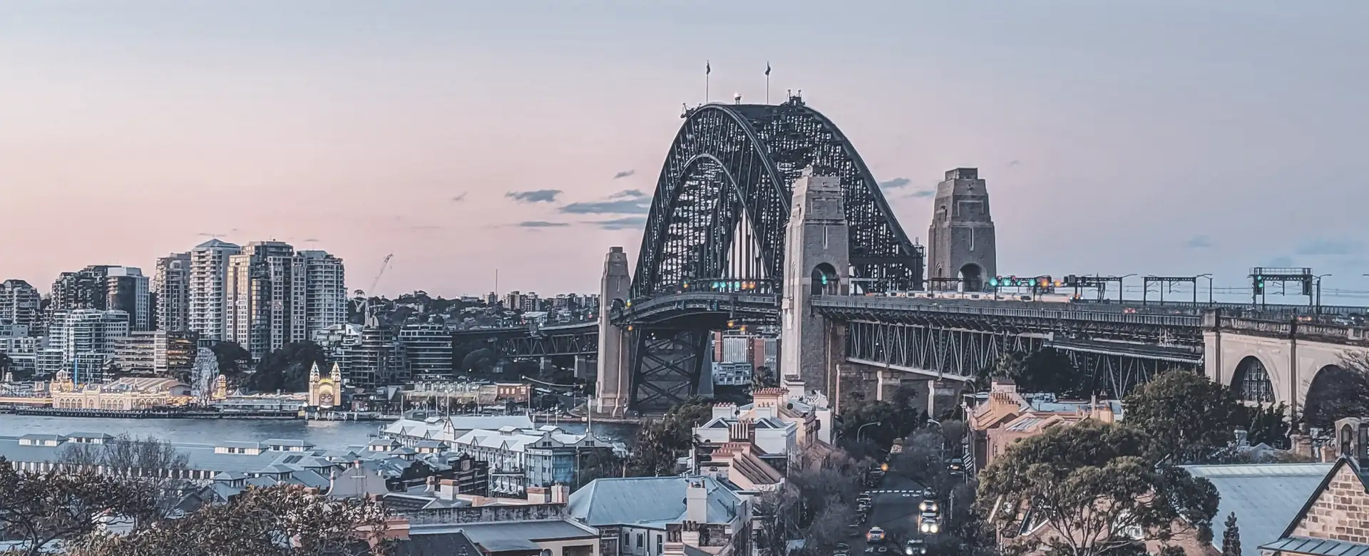 A harbour bridge and city towers at dusk, seen over rooftops